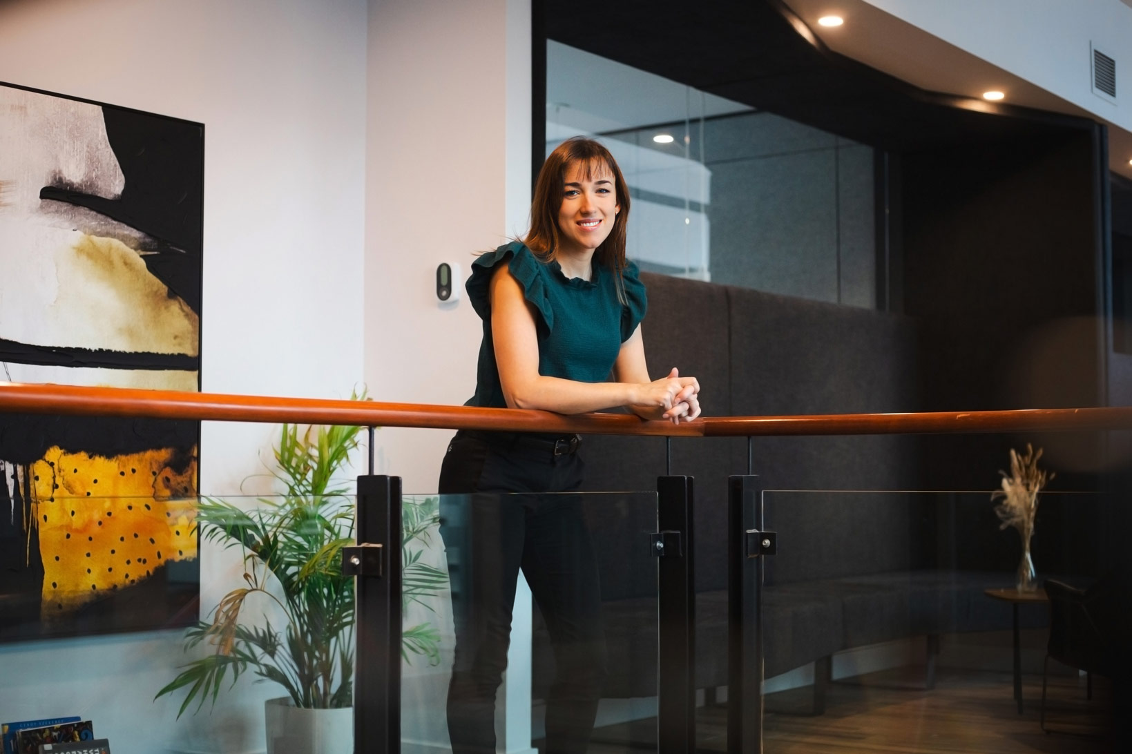Une femme souriante se tient près d'une balustrade dans un environnement moderne. Elle porte un haut vert et un pantalon noir, avec un tableau coloré en arrière-plan.